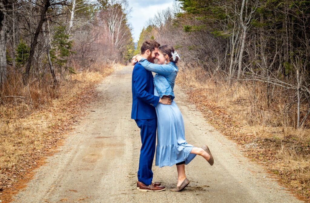 This is a header image showing a woman in a blue dress hugging a man in a blue suit, standing on a dirt trail in the woods during autumn.